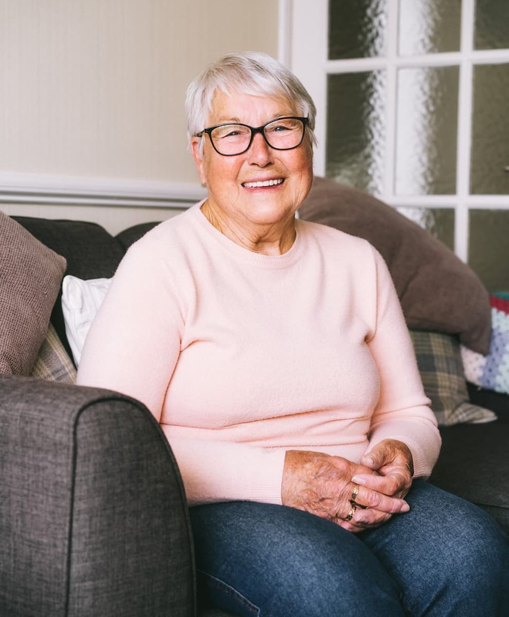 Femme âgée à lunettes souriant, assise sur un canapé, mains jointes